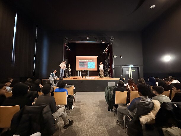 Voll besetzte Aula, Schülerpublikum blickt zur Bühne mit Präsentation