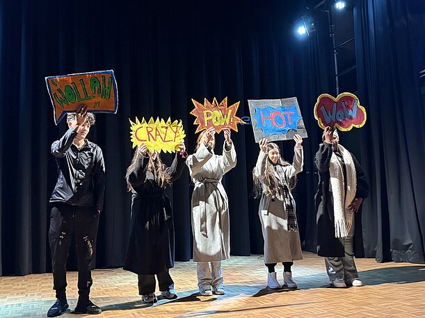Nahaufnahme: Theatergruppe mit Schildern „Crazy“, „Pow“ und „Hot“ vor dunklem Vorhang