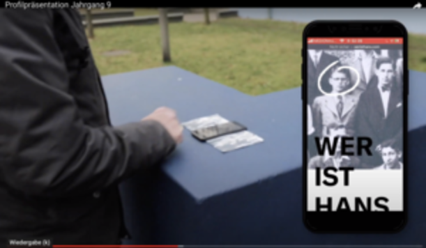 Smartphone mit Wer ist Hans neben alten Fotos auf blauem Sockel