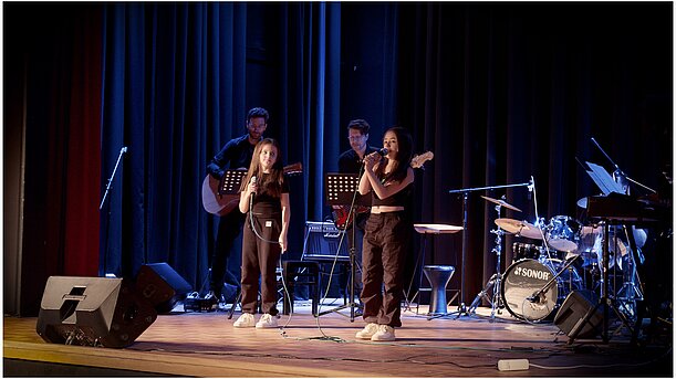 Zwei Sängerinnen im Duett vor Schlagzeug und Band auf dunkler Bühne