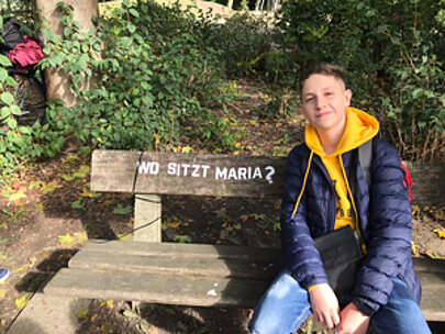 Schüler sitzt auf einer Holzbank mit der Aufschrift Wo sitzt Maria