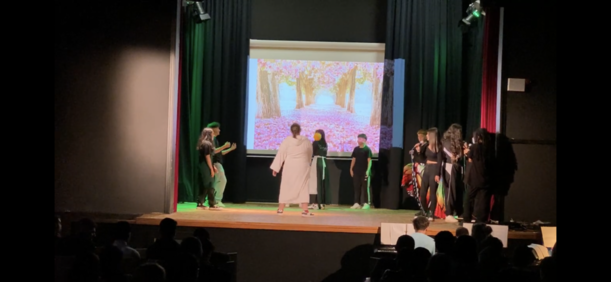 Mehrere Darsteller in Kostümen vor projiziertem Zauberwald im Theaterstück