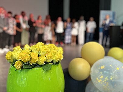 Gelbe Rosen in grüner Vase und Ballons, dahinter Absolventen auf der Bühne