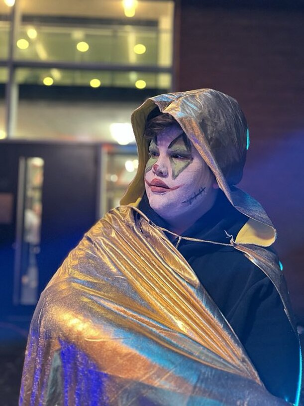 Person mit silbrig-goldenem Umhang und weiß geschminktem Gesicht bei Nacht