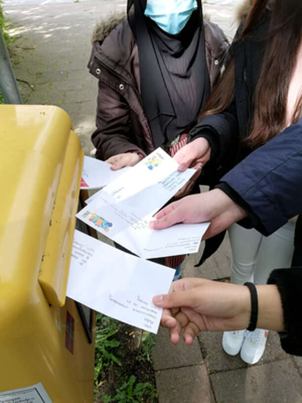Hände stecken mehrere Briefumschläge in einen gelben Briefkasten