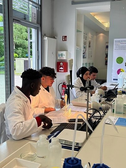 Schülerinnen und Schüler in Laborkitteln an Arbeitsplätzen im Labor