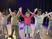 Kinder mit roten Clownsnasen und bunten Hüten strecken auf der Bühne die Arme hoch