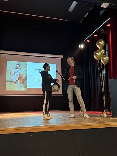 Zwei Personen mit Mikrofon auf der Bühne vor Leinwand mit Foto