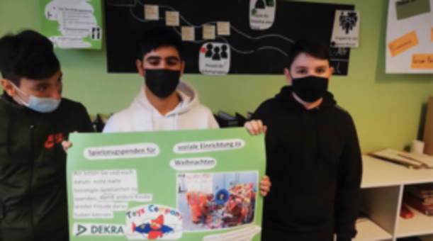 Drei Schüler mit Masken zeigen ein Plakat zu Spielzeugspenden für Weihnachten