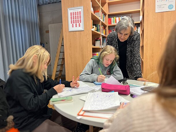 Kinder schreiben in Hefte, eine Lehrerin schaut ihnen über die Schulter