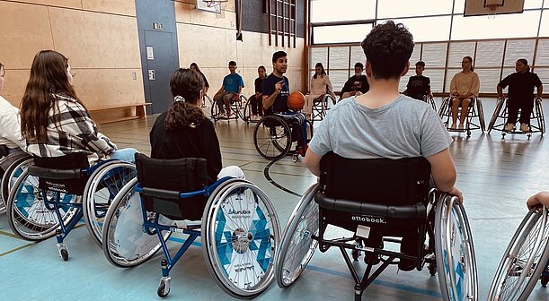 Spieler der BG Baskets erklärt Ballmitnahme im Rollstuhl, Jugendliche schauen zu