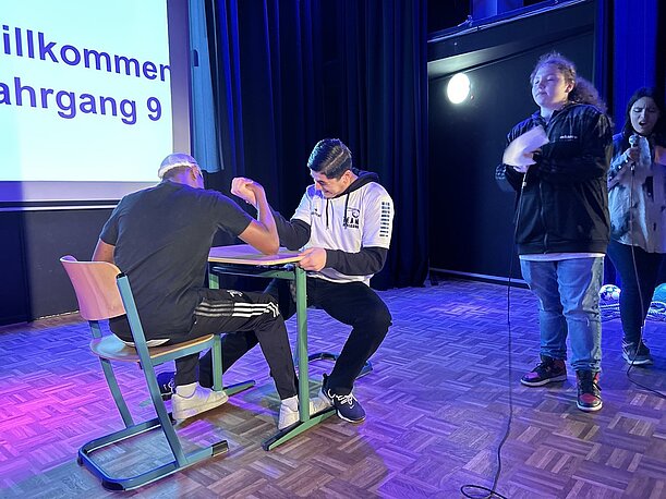 Szene auf der Bühne: zwei Schüler beim Armdrücken am Schultisch
