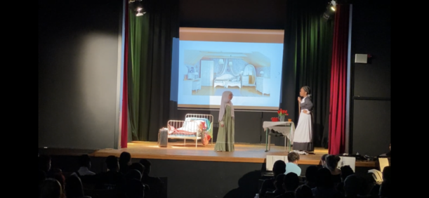 Darstellerinnen neben Bett vor projiziertem Zimmer im Theaterstück der 8d