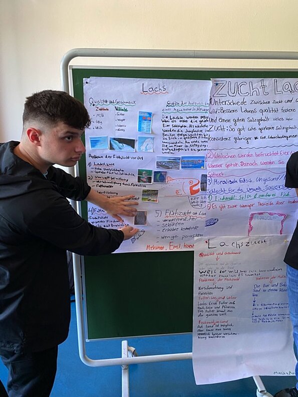 Anid zeigt auf ein Plakat zum Thema Lachszucht