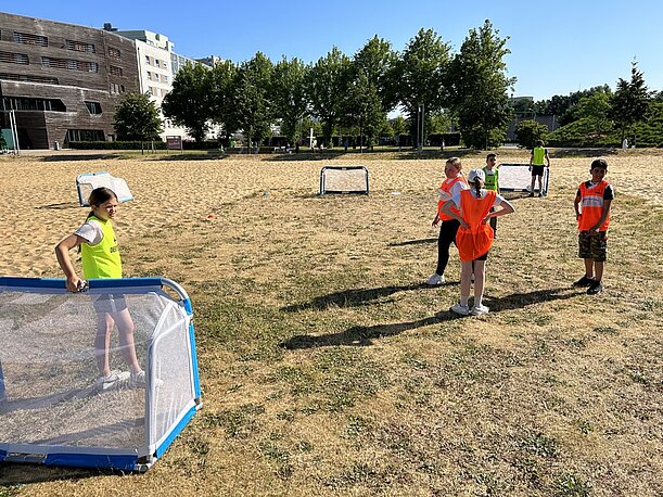 Kinder in Leibchen spielen mit kleinen Toren auf einem Sandplatz
